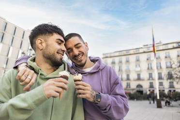 Happy gay couple with arms around holding ice cream at city street - JCCMF09154