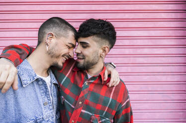 Gay couple standing with arms around each other in front of roller shutter wall - JCCMF09160