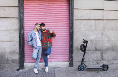 Happy gay couple standing in front of roller shutter by electric push scooter - JCCMF09164