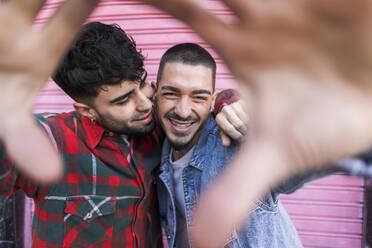 Happy gay couple gesturing in front of pink roller shutter - JCCMF09165