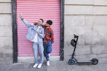 Young gay couple taking selfie using smart phone in front of roller shutter wall - JCCMF09167