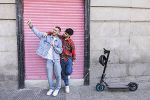 Junges schwules Paar macht Selfie mit Smartphone vor einer Rollladenwand - JCCMF09167