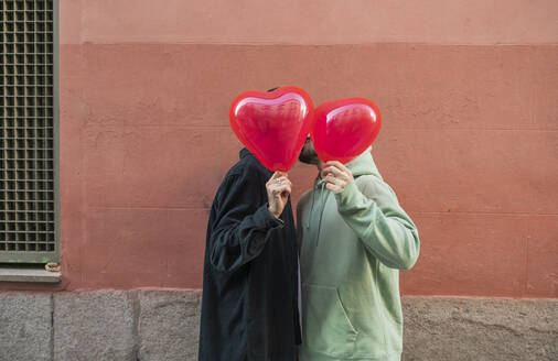 Gay couple covering face with red heart shape balloon - JCCMF09199