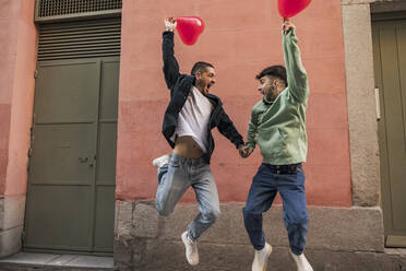 Glückliches homosexuelles Paar, das mit roten herzförmigen Luftballons springt - JCCMF09206