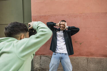 Man clicking photos of friend gesturing in front of wall - JCCMF09211