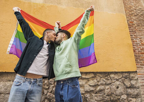 Homosexuelles Paar mit Regenbogenflagge küsst sich vor einer Mauer - JCCMF09213