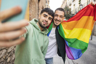 Glückliches schwules Paar mit Regenbogenfahne, das ein Selfie mit seinem Smartphone auf dem Fußweg macht - JCCMF09216