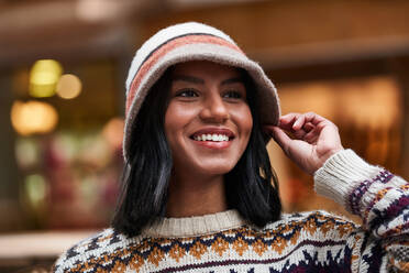 Optimistic young black female in knitted sweater and stylish hat smiling on blurred background of building on city street - ADSF42864