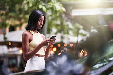 Junge schwarze Frau in stilvoller Kleidung, die eine SMS auf einem Smartphone auf einem unscharfen Hintergrund eines Parks an einem Sommerwochenende schreibt - ADSF42865