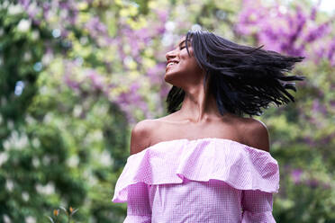Young black woman shaking hair while standing near lush tree with small purple flowers in park - ADSF42867