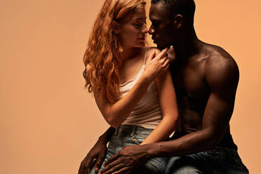 Rothaarige Freundin im Top und hemdsärmeliger afroamerikanischer Freund umarmen sich zärtlich und schauen sich an, während sie im Studio sitzen - ADSF42870