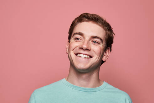 Positiver junger Rothaariger in schwarzem T-Shirt, der mit einem Lächeln wegschaut, während er vor einem rosa Hintergrund steht - ADSF42879