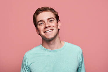 Positiver junger Rothaariger in schwarzem T-Shirt, der lächelnd in die Kamera schaut, während er vor einem rosa Hintergrund steht - ADSF42881