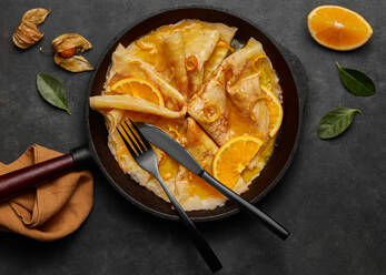 Von oben hausgemachte Crêpes Suzette mit Orangenlikör und Orangenscheiben, serviert auf einem Teller mit Messer und Gabel auf einer Betonfläche - ADSF42895