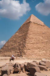 Seitenansicht eines nicht erkennbaren Beduinen auf einem Kamel in der Nähe der antiken Pyramide von Gizeh an einem sonnigen Tag in der Wüste in Ägypten - ADSF42899