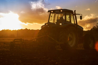 Seitenansicht eines nicht erkennbaren Landwirts, der einen Traktor fährt, während er den Boden eines landwirtschaftlichen Feldes unter bewölktem Himmel an einem sonnigen Tag bearbeitet - ADSF42942