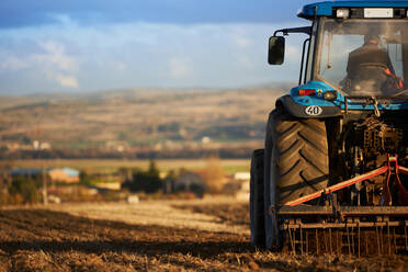 Rückansicht eines nicht erkennbaren Landwirts, der einen Traktor fährt, während er den Boden eines landwirtschaftlichen Feldes unter bewölktem Himmel an einem sonnigen Tag bearbeitet - ADSF42943