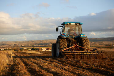 Rückansicht eines nicht erkennbaren Landwirts, der einen Traktor fährt, während er den Boden eines landwirtschaftlichen Feldes unter bewölktem Himmel an einem sonnigen Tag bearbeitet - ADSF42944