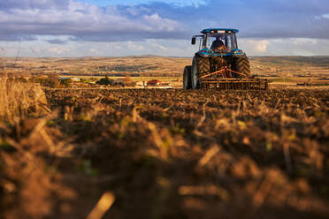 Rückansicht eines nicht erkennbaren Landwirts, der einen Traktor fährt, während er den Boden eines landwirtschaftlichen Feldes unter bewölktem Himmel an einem sonnigen Tag bearbeitet - ADSF42945