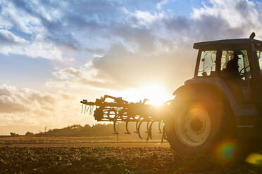 Seitenansicht eines nicht erkennbaren Landwirts, der einen Traktor fährt, während er den Boden eines landwirtschaftlichen Feldes unter bewölktem Himmel an einem sonnigen Tag bearbeitet - ADSF42946