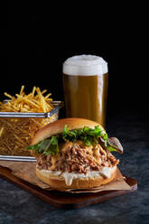 Appetitlicher Burger mit Pulled Pork mit Barbecue-Sauce, Cheddar-Käse und Salatmischung, serviert mit Pommes Frites und einem Glas kaltem Bier auf einem Holzbrett - ADSF42947