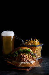 Appetitlicher Burger mit Pulled Pork mit Barbecue-Sauce, Cheddar-Käse und Salatmischung, serviert mit Pommes Frites und einem Glas kaltem Bier auf einem Holzbrett - ADSF42948