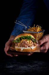 Crop anonyme Person mit appetitlichen Burger mit frischem Salat Schweinefleisch und Französisch Brände auf dunklem Hintergrund - ADSF42949