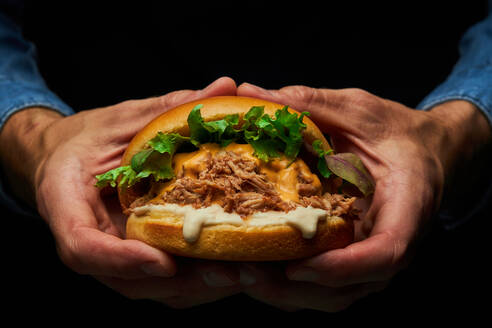 Crop anonyme Person mit appetitlichen Burger mit frischem Salat Schweinefleisch und Käse auf dunklem Hintergrund - ADSF42950