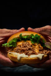 Crop anonyme Person mit appetitlichen Burger mit frischem Salat Schweinefleisch und Käse auf dunklem Hintergrund - ADSF42951