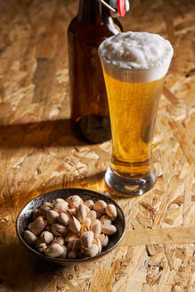 From below glass of fresh cold beer placed on wooden table near bottle and plate with pistachios - ADSF42952