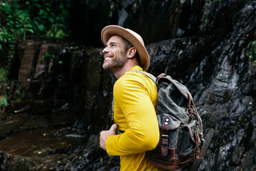 Seitenansicht eines männlichen Touristen mit Hut und gelbem Pullover, der einen Rucksack trägt und die malerische Landschaft bewundert - ADSF42960