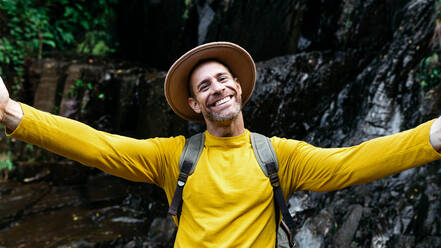 Glücklicher männlicher Tourist mit Hut und gelbem Pullover, der einen Rucksack trägt und die malerische Landschaft bewundert - ADSF42962