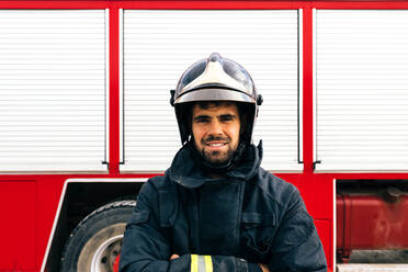 Positive Hispanic fireman in protective uniform with arms crosses looking at camera with smile standing near fire engine on station - ADSF42963