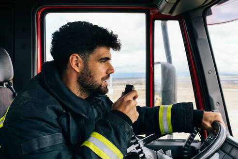 Seitenansicht eines bärtigen hispanischen Feuerwehrmanns in Uniform, der wegschaut und in ein Funkgerät spricht, während er ein Feuerwehrauto zu einem Notfallort steuert - ADSF42981