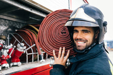 Seitenansicht eines fröhlichen hispanischen Feuerwehrmannes in Uniform und mit Helm, der einen Schlauch auf der Schulter trägt und in die Kamera schaut, in der Nähe eines Feuerwehrautos bei Tageslicht - ADSF42982