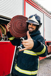 Fröhlicher hispanischer Feuerwehrmann in Uniform und Helm, der einen Schlauch auf der Schulter trägt und ein Shaka-Zeichen macht, während er in der Nähe eines Löschfahrzeugs steht - ADSF42983