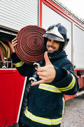 Happy Hispanic firefighter in uniform and helmet carrying hose on shoulder and gesturing shaka sign while standing near fire engine in daytime - ADSF42983