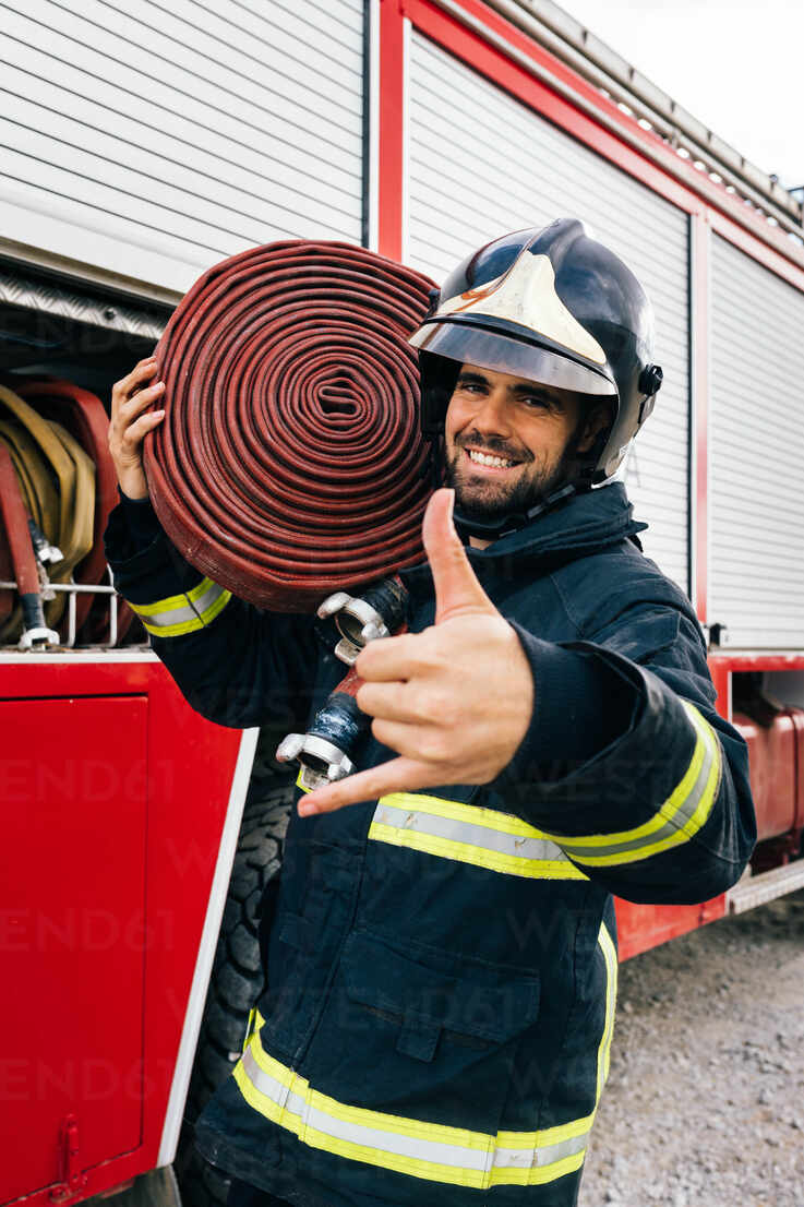 Happy Hispanic firefighter in uniform and helmet carrying hose on shoulder  and gesturing shaka sign while standing near fire engine in daytime stock  photo