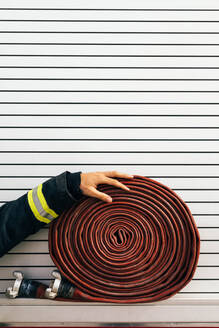 Anonymer Feuerwehrmann in Arbeitsschutzkleidung, der einen aufgerollten Schlauch berührt, der an der Seite eines Feuerwehrfahrzeugs angebracht ist - ADSF42984