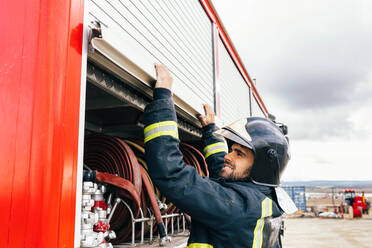 Positive focused Hispanic firefighter smiling and closing hose compartment of fire engine against cloudy sky on station - ADSF42985