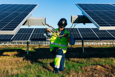 Arbeiter in Uniform und mit Schutzhelm beim Anschließen von Drähten bei der Installation von Photovoltaikzellen auf einem Feld an einem sonnigen Tag - ADSF42986