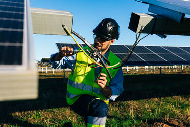 Arbeiter in Uniform und mit Schutzhelm beim Anschließen von Drähten bei der Installation von Photovoltaikzellen auf einem Feld an einem sonnigen Tag - ADSF42987