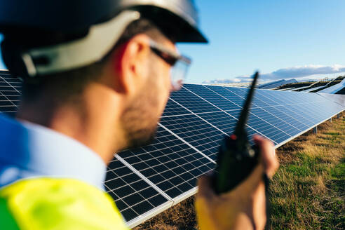 Rückenansicht eines unscharfen anonymen Technikers, der sich mit einem Walkie-Talkie über die in der Landschaft platzierten Fotovoltaikzellen bei Sonnenschein meldet - ADSF42989
