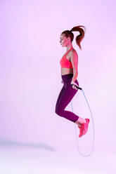 Side view of fit female athlete with ponytail jumping with rope during fitness training against lilac background - ADSF43046
