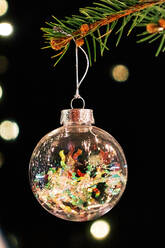 Nahaufnahme einer transparenten Glaskugel mit buntem Konfetti, die an einem Zweig eines Weihnachtsbaums in einem dunklen Raum mit leuchtenden Lichtern hängt - ADSF43053