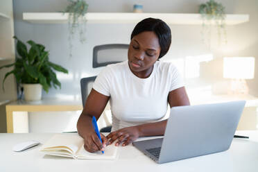 Junge schwarze Frau in weißem T-Shirt schreibt Daten in einen Planer in der Nähe eines Laptops, während sie zu Hause am Tisch sitzt und lernt - ADSF43063