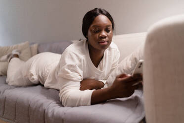 Young black female in white pajama lying on coach with soft pillow and browsing social media on smartphone in living room in evening at home - ADSF43064