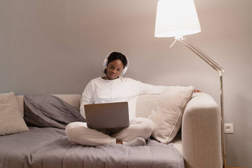 Young African American female in white pajama and socks with headphones browsing movie on laptop while sitting cross legged on couch in living room at home - ADSF43065