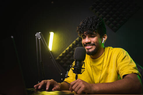 Junger hispanischer Mann in gelbem T-Shirt mit TWS-Kopfhörern lächelt in der Nähe des Mikrofons, während er an einem Schreibtisch in einem dunklen Studio mit Neonbeleuchtung sitzt und einen Podcast aufnimmt - ADSF43069