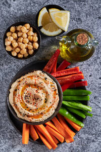 Draufsicht auf köstlichen Hummus und verschiedene Gemüsescheiben, serviert neben einer Flasche Öl und Schalen mit Kichererbsen- und Zitronenstücken auf einem grauen Marmortisch - ADSF43082